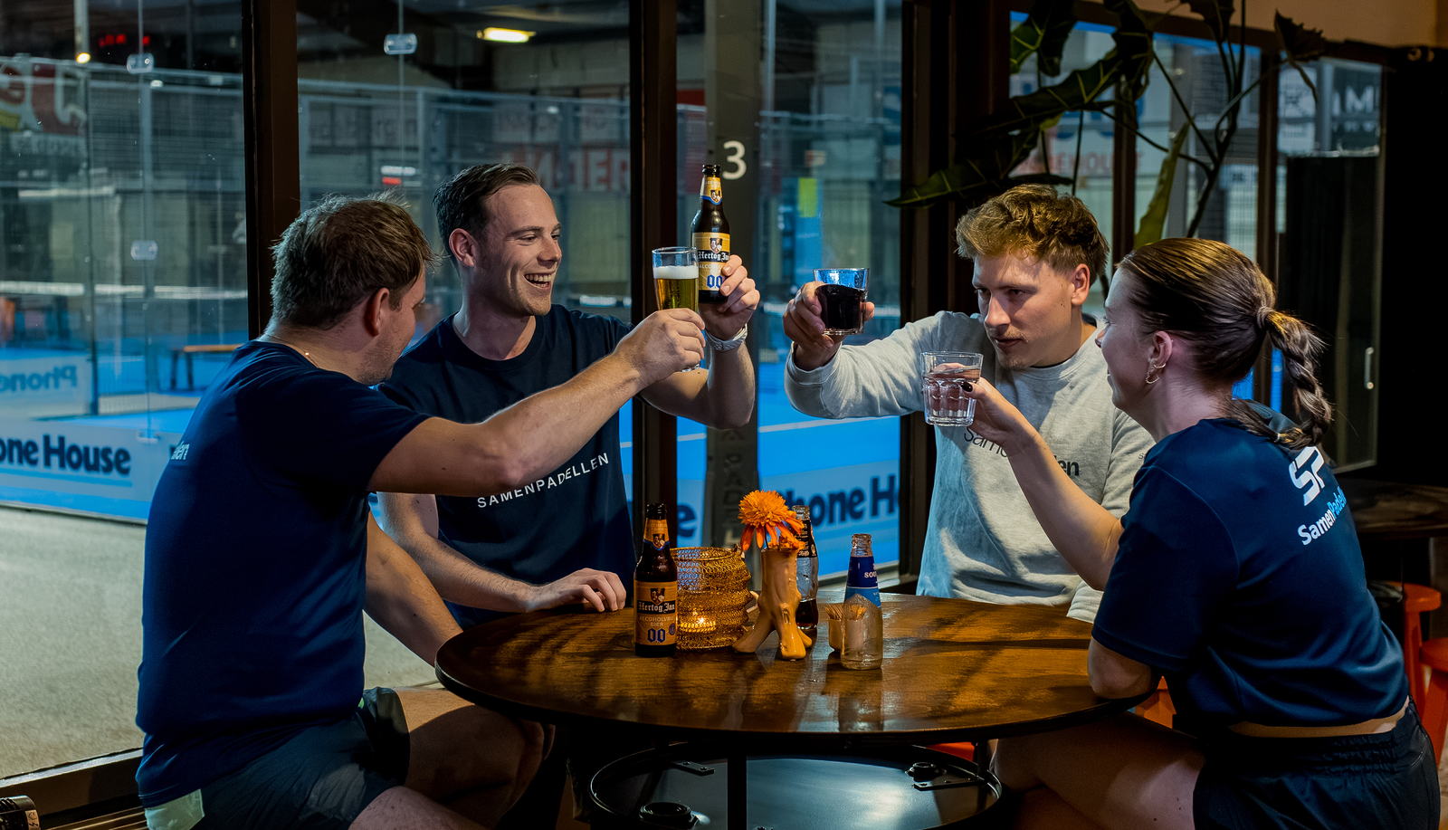 Vier spelers proosten na afloop, met de padelbaan achter het glas